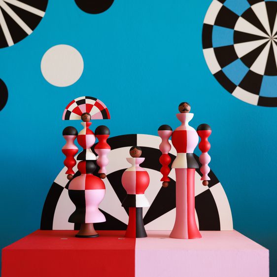 A vibrant display of three abstract wooden sculptures in red, pink, and black, set against a bright blue wall adorned with geometric patterns and circular designs in black, white, and blue. The sculptures feature varied shapes and heights, creating a playful aesthetic.