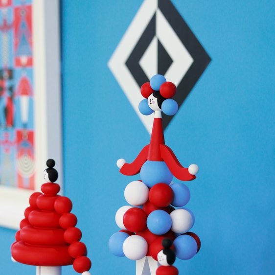 A vibrant display of abstract sculptures in red, white, and blue, featuring rounded shapes and playful designs. The background is a bright blue wall with geometric patterns, complemented by a framed artwork featuring similar colours and shapes.
