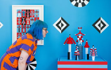 A woman with blue hair leans towards a display of colourful, abstract sculptures on a table. The background features a vibrant blue wall adorned with geometric patterns in red, black, and white, alongside a framed artwork.
