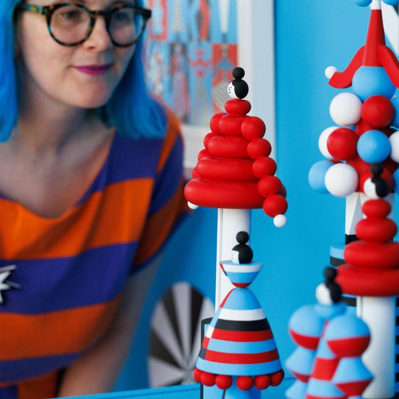 A woman with blue hair and glasses admires colourful sculptures resembling stylised figures and trees. The display features red, white, and blue elements, set against a vibrant blue background with geometric patterns.