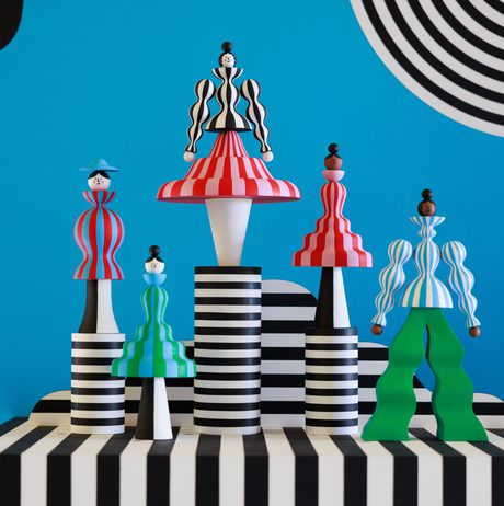 A vibrant display of stylised figurines in bold colours and patterns, set against a bright blue wall with black and white stripes. The figures feature exaggerated shapes and playful designs, creating a striking visual contrast.
