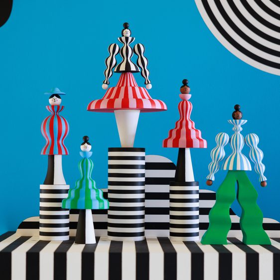 A vibrant display of stylised figurines in bold colours and patterns, set against a bright blue wall with black and white stripes. The figures feature exaggerated shapes and playful designs, creating a striking visual contrast.