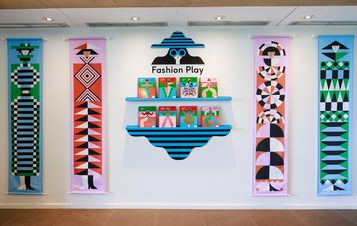 A display of four colourful graphic wall hangings against a white wall. In the centre is a set of shelves filled with copies of the book Fashion Play.