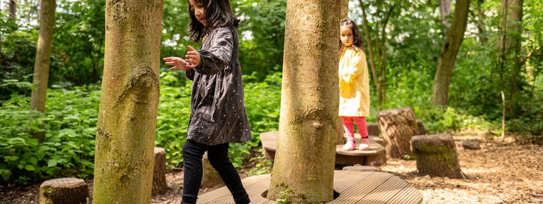Two children jumping between wooden platforms in a nature play area.