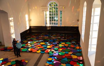 An adult and child using a mirrored disc to cast reflections on a wall in a gallery space, filled with a rainbow of coloured discs installed at floor level
