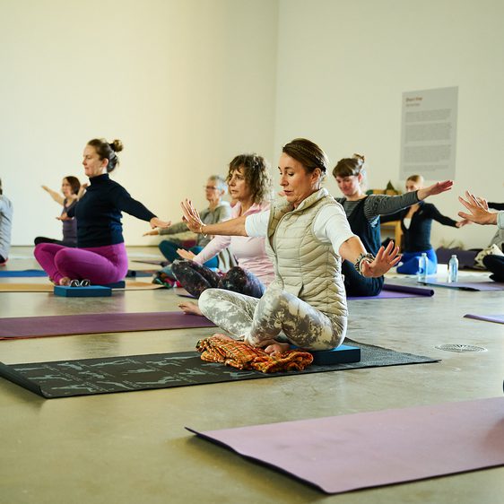A group of people sitting on yoga mats and practicing yoga inside a gallery space.