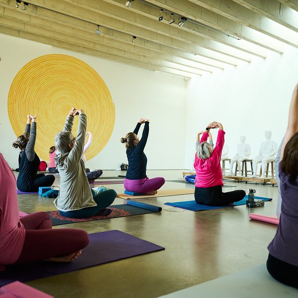 A group of people practicing yoga inside a gallery space. A yellow circular artwork on a wall and a row of plaster figures are in the background.