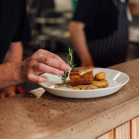 A chef adds a pea shoot garnish to a plate of roasted belly pork and vegetables resting on a stone counter top.