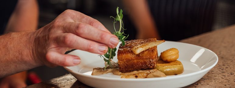 A chef adds a pea shoot garnish to a plate of roasted belly pork and vegetables resting on a stone counter top.