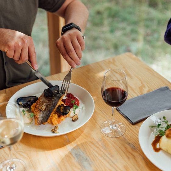 A person cuts in to a piece of grilled fish on a white plate. Glasses of wine, napkins and cutlery are on the table around them.