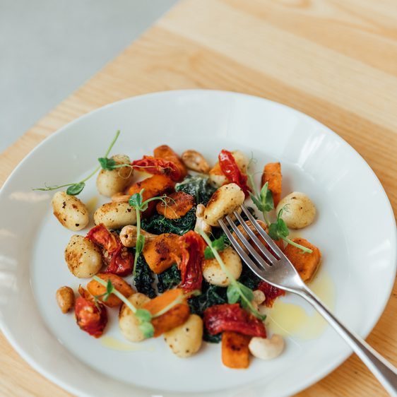 A plate of gnocci and roasted vegetables on a wooden table, with a fork above the dish.