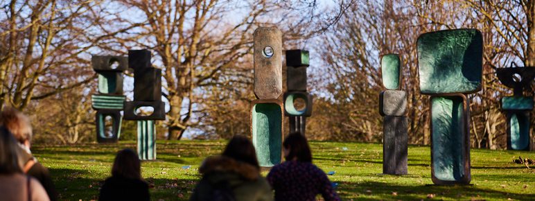 A group of people walking towards a display of tall bronze abstract sculptures displayed outdoors.