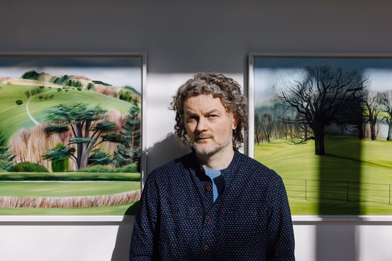 A man with curly grey hair, wearing a nay jumper, standing in front of paintings of landscapes.