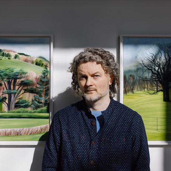 A man with curly grey hair, wearing a nay jumper, standing in front of paintings of landscapes.