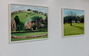 Two landscape paintings depicting tress and hills displayed on a gallery wall.