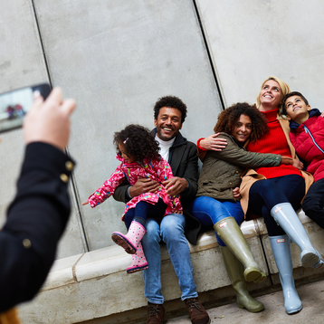 A group of people sitting on a low concrete bench with tall walls behind it, having their photo taken by a person with a mobile phone.