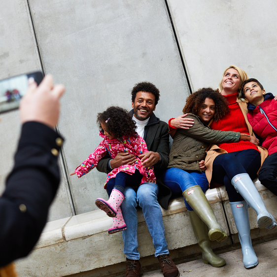 A group of people sitting on a low concrete bench with tall walls behind it, having their photo taken by a person with a mobile phone.
