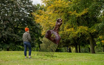 A woman wearing a coat and wooly hat looking at a sculpture of an oversized afro pick with a black power as a handle, displayed outdoors.