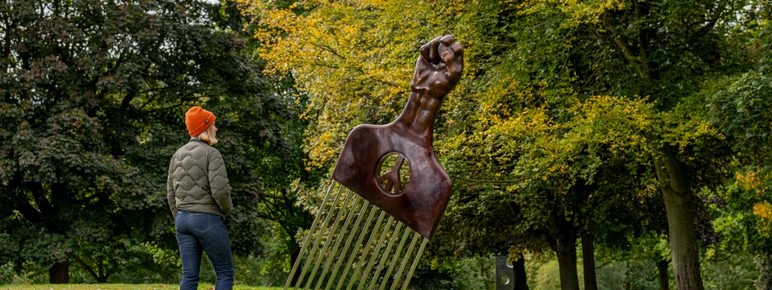 A woman wearing a coat and wooly hat looking at a sculpture of an oversized afro pick with a black power as a handle, displayed outdoors.