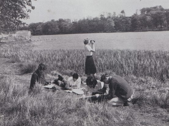 A black and white archive photo of a group of people working next to a lake. One is standing and looking across the lake with binoculars.