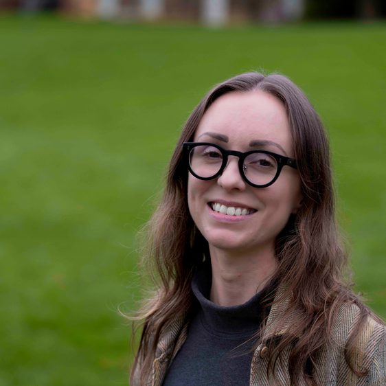 A woman with shoulder length brown hair and glasses standing outdoors.
