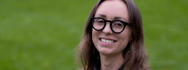 A woman with shoulder length brown hair and glasses standing outdoors.