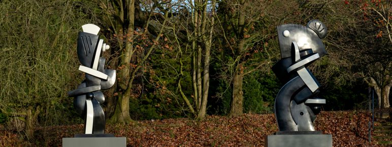Two black and white abstract sculptures displayed on plinths outdoors, with trees behind them.