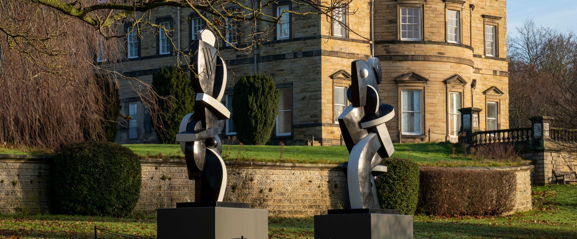 Two black and white abstract sculptures displayed on plinths outdoors. A historic manor house is behind them.