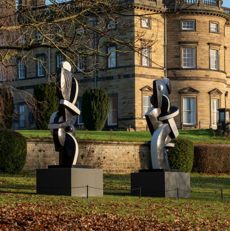 Two black and white abstract sculptures displayed on plinths outdoors. A historic manor house is behind them.