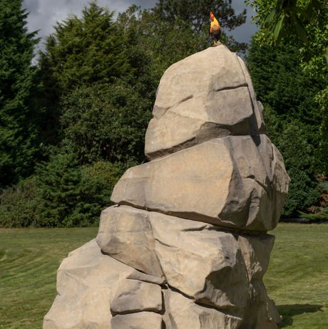 A sculpture depicting a stack of boulders with a life-sized chicken perched on the top, displayed outdoors.