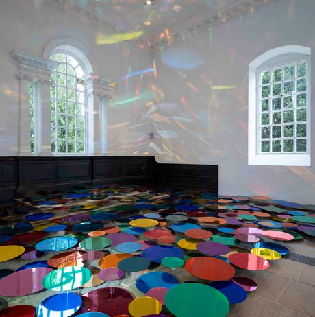 An installation of colourful mirrored discs in a Chapel space with tall windows. The discs are displayed at different heights to cast reflections on the walls.