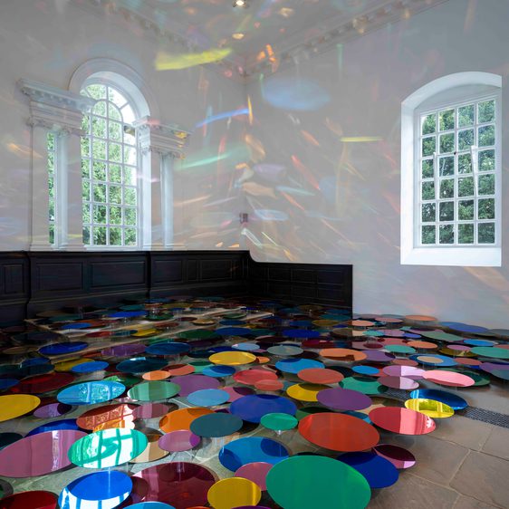 An installation of colourful mirrored discs in a Chapel space with tall windows. The discs are displayed at different heights to cast reflections on the walls.