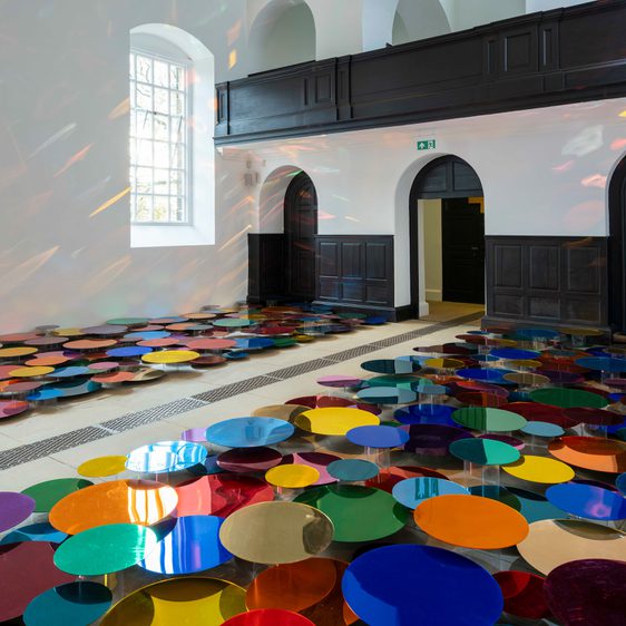 An installation of colourful mirrored discs in a Chapel space with tall windows. The discs are displayed at different heights to cast reflections on the walls.
