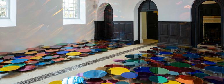 An installation of colourful mirrored discs in a Chapel space with tall windows. The discs are displayed at different heights to cast reflections on the walls.