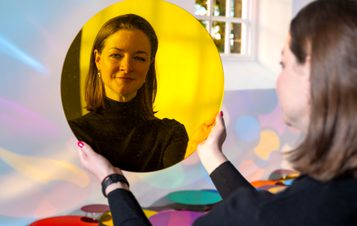 A woman holding up a yellow mirrored disc and looking at her reflection.
