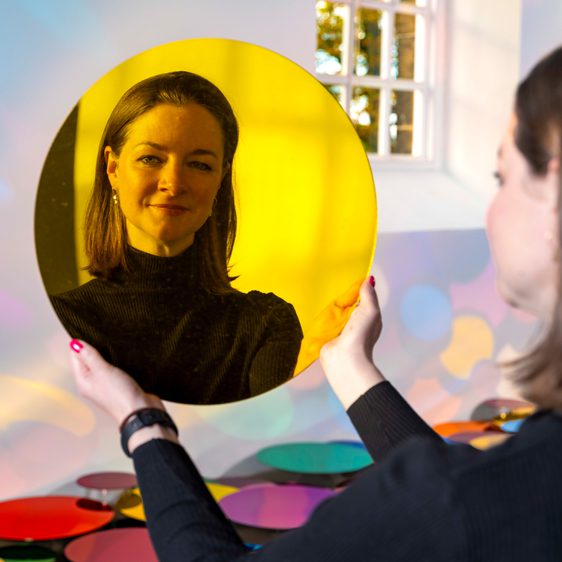A woman holding up a yellow mirrored disc and looking at her reflection.