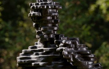 Detail of a sculpture of a human figure made from layers of bronze, displayed outdoors with foliage behind it.