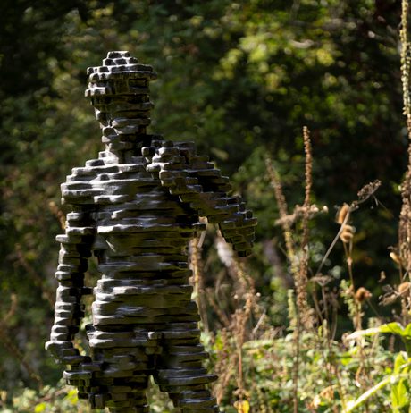 A sculpture of a human figure made from layers of bronze, displayed on a concrete plinth outdoors with foliage behind it.