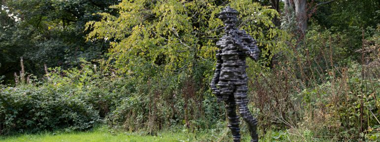 A sculpture of a human figure made from layers of bronze, displayed on a concrete plinth outdoors with foliage behind it.