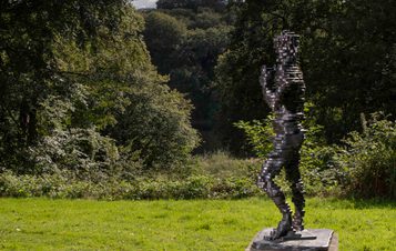 A sculpture of a human figure made from layers of bronze, displayed on a concrete plinth outdoors with foliage behind it.
