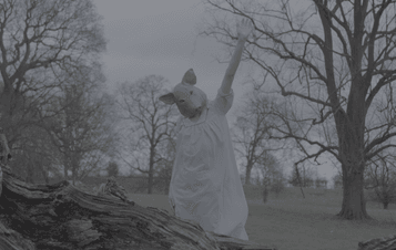 A woman wearing a pig mask and blue nightgown, standing outdoors between trees with one arm in the air.