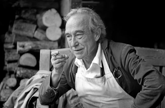 A thoughtful older man sits on a wooden bench, wearing an apron over a white shirt. He holds a cigarette and gazes into the distance, with a backdrop of stacked logs. The image is in black and white, highlighting his contemplative expression.