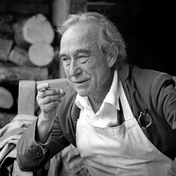 A thoughtful older man sits on a wooden bench, wearing an apron over a white shirt. He holds a cigarette and gazes into the distance, with a backdrop of stacked logs. The image is in black and white, highlighting his contemplative expression.