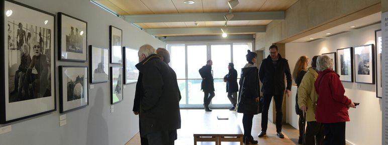 A group of visitors explores a photography exhibition in a well-lit gallery. Black and white photographs hang on the walls, while people of various ages engage with the artwork, some reading descriptions and others discussing the images.