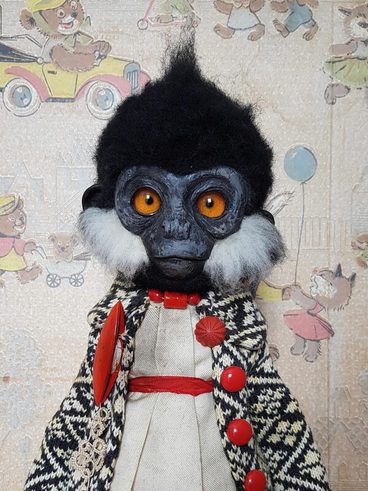 A vintage-style doll resembling a monkey, featuring large orange eyes, a black tufted hairstyle, and white facial fur. It wears a patterned coat with red buttons over a cream dress, set against a whimsical, illustrated background.
