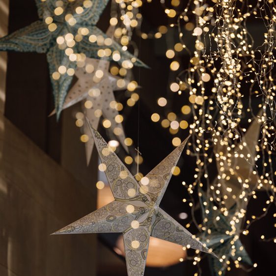 A close-up of decorative paper stars hanging from the ceiling, surrounded by twinkling fairy lights. The stars are intricately patterned, adding a festive touch to the warm, softly lit atmosphere.