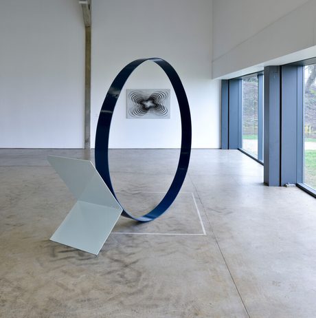 A spacious gallery features three contemporary sculptures: a red zigzag structure, a large blue circular form, and a sleek white angular piece. A black and white abstract artwork hangs on the wall, with large windows revealing a green landscape outside.