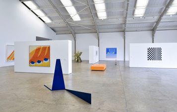 A spacious art gallery featuring various contemporary artworks. On the walls, there are colourful paintings and geometric designs. In the centre, a blue sculpture and an orange rectangular platform add to the modern aesthetic of the space.