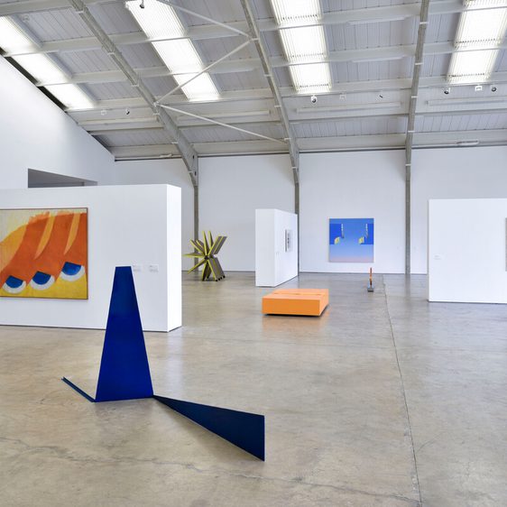 A spacious art gallery featuring various contemporary artworks. On the walls, there are colourful paintings and geometric designs. In the centre, a blue sculpture and an orange rectangular platform add to the modern aesthetic of the space.