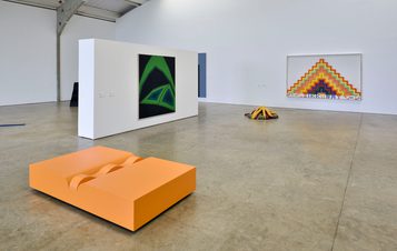 A spacious art gallery featuring three abstract artworks. On the left, a large green and black painting; in the centre, an orange sculptural bench; and on the right, a colourful geometric piece with layered shapes. The floor is polished concrete.